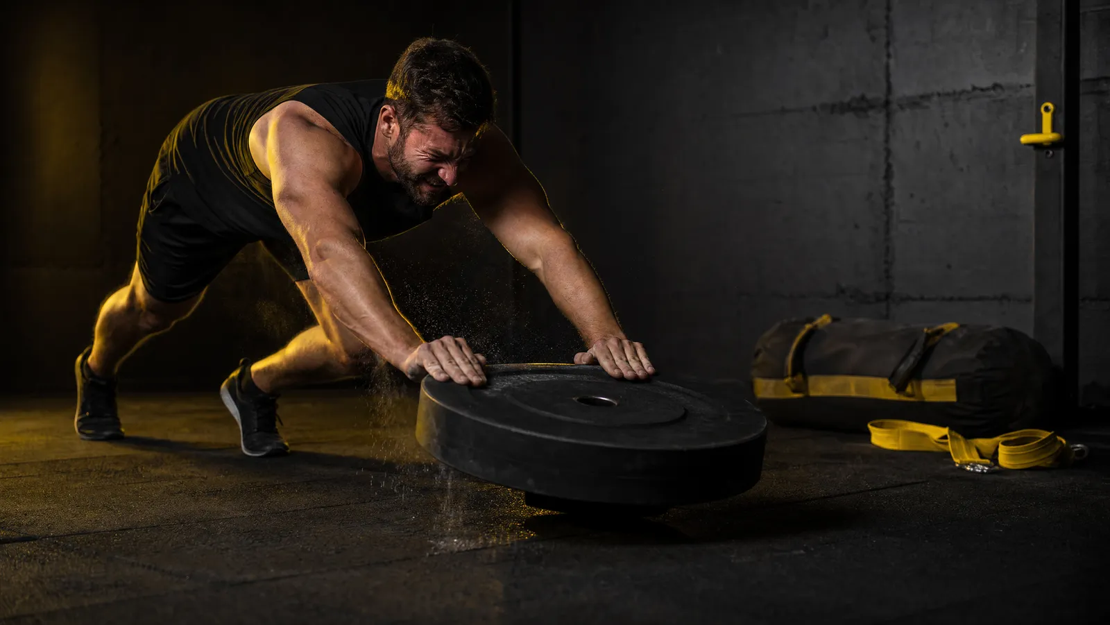 Athlete pushing a heavy weight plate across a rubber floor in a dark home garage gym