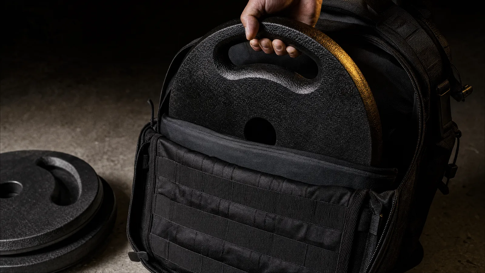 Cast iron ruck plate with a grab handle being lowered into the padded back sleeve of an open tactical backpack on a garage floor